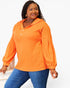 A woman modeling a bright orange long-sleeve pullover top with a buttoned collar and puff sleeves, paired with blue jeans, against a plain white studio background.