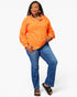 A woman modeling a bright orange long-sleeve hooded top with blue jeans and black slide sandals, standing against a light gray studio background.