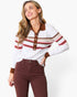 A woman wearing a white long-sleeve knit polo-style sweater with brown and red striped accents across the chest and sleeves, paired with dark brown pants, against a plain white studio background.