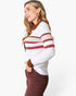 A woman wearing a white ribbed knit polo-style sweater with brown, red, and dark brown horizontal stripes on the chest and sleeves, paired with brown pants, photographed against a clean white studio background.