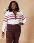 Woman wearing a white sweater with brown and red stripes and brown pants against a beige background