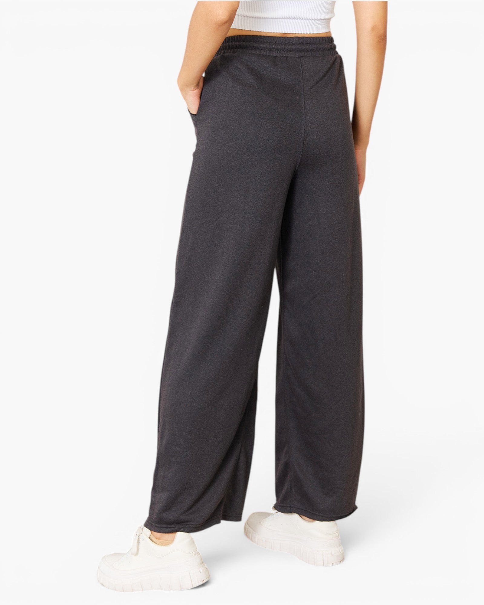A person wearing dark charcoal wide-leg sweatpants with a drawstring waist, paired with a white crop top and white sneakers, shown from the waist down on a light gray studio background.