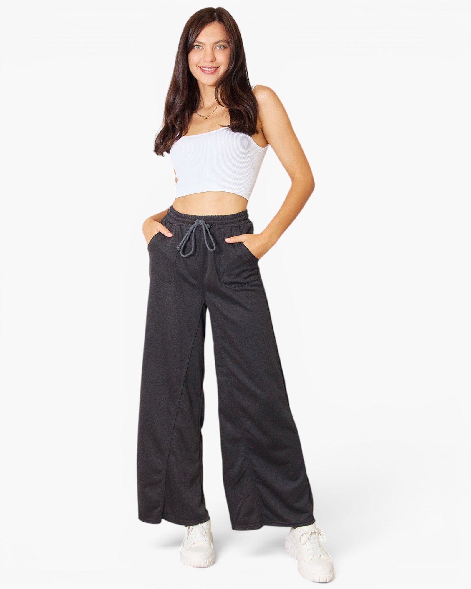 A woman modeling a pair of wide-leg charcoal gray drawstring pants with a white strapless crop top and white sneakers, standing against a solid white studio background.