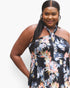 A woman wearing a halter-neck floral dress with a black base and multicolor watercolor-style flowers in blue, pink, and peach, photographed against a plain light gray background.