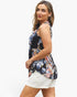 A woman modeling a sleeveless floral top with a navy base and multicolor botanical print, paired with white shorts, against a plain white studio background.