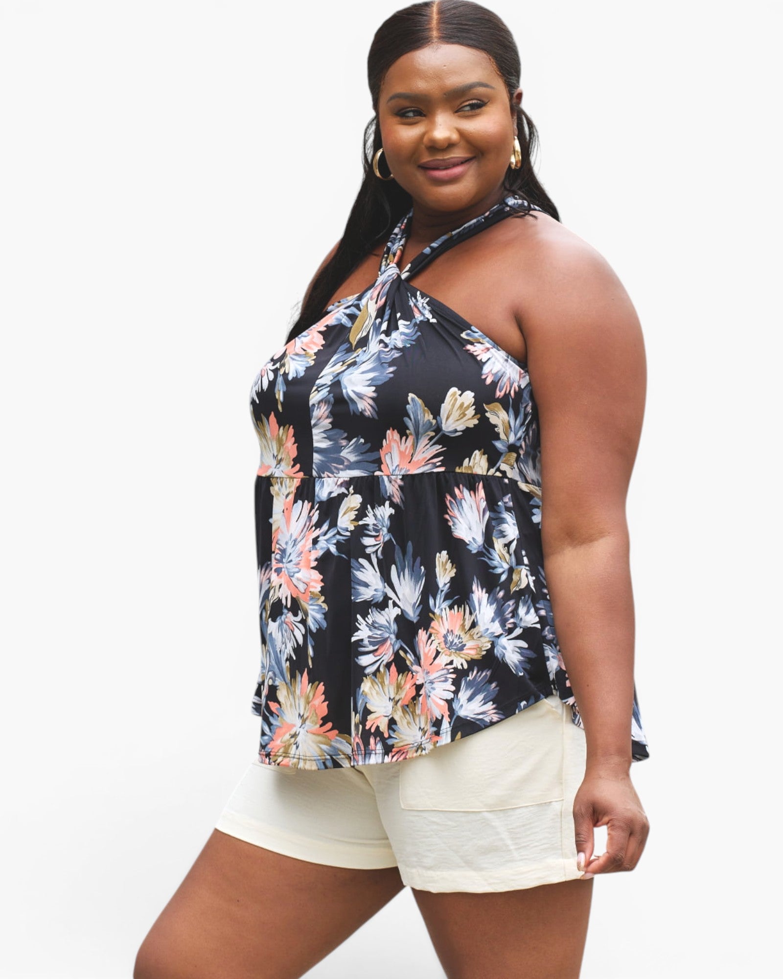 A plus-size model wearing a black halter tank top with a colorful floral print and cream shorts, photographed against a solid white studio background.
