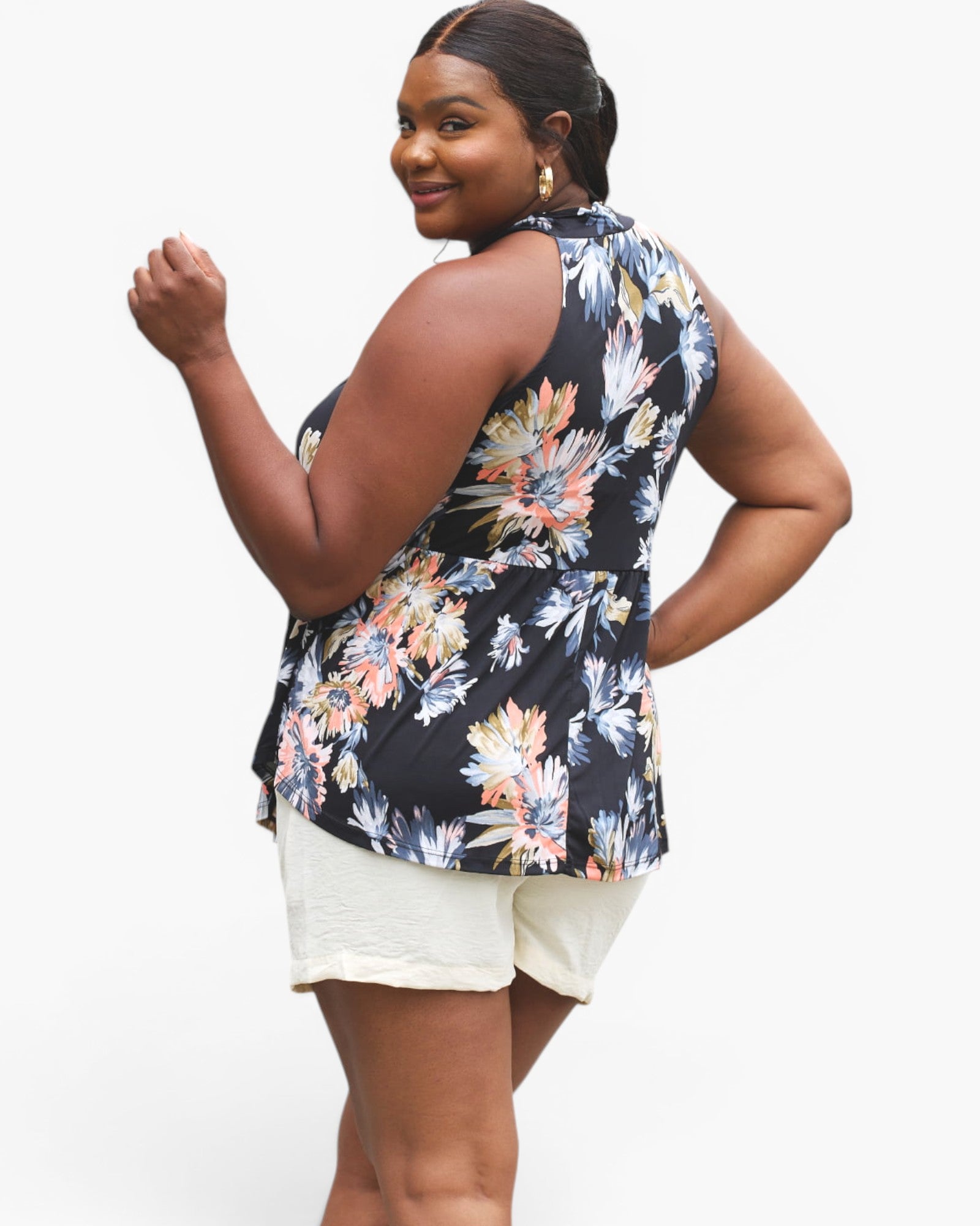 A woman modeling a sleeveless floral top with a black base and multicolor floral print, paired with cream shorts, against a light gray studio background.