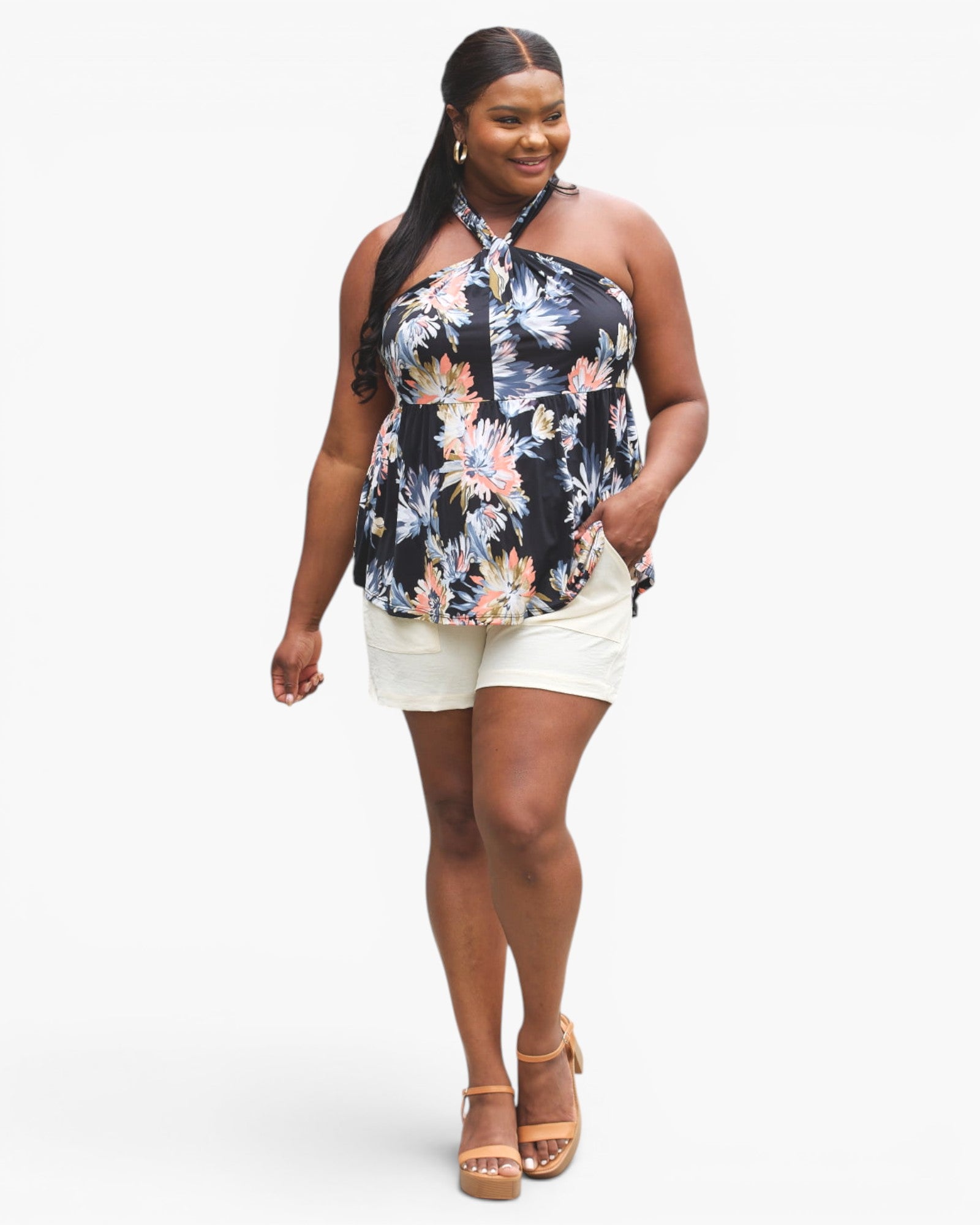 A woman modeling a plus-size halter top with a black base and a colorful floral print in pink, blue, and white, paired with white shorts and tan platform sandals, against a light gray studio background.