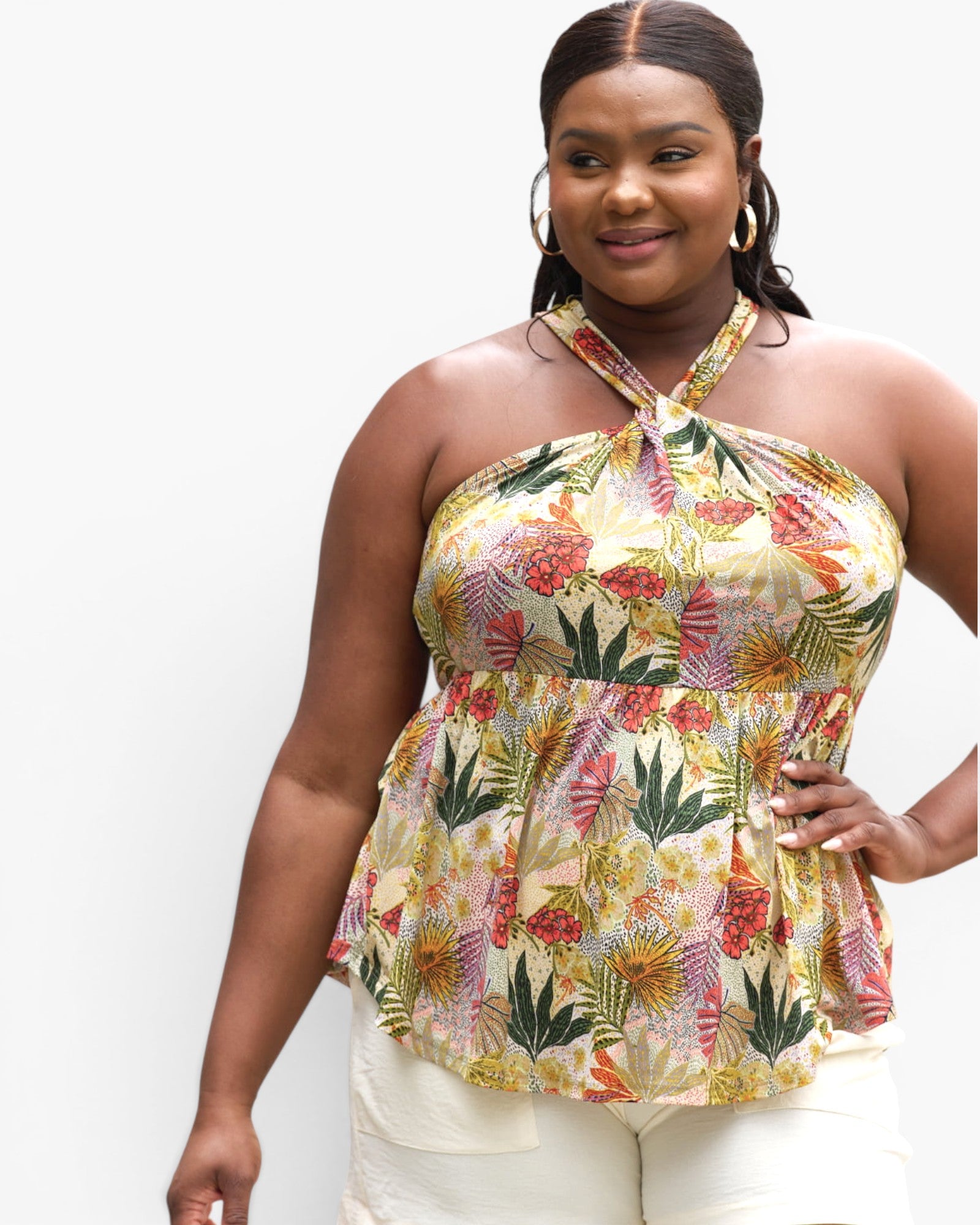 A woman modeling a halter-neck peplum top with a colorful tropical floral and leaf print on a light cream background, paired with white pants, against a plain white studio background.