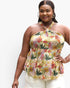 A woman modeling a halter-neck peplum top with a colorful tropical floral and leaf print on a light cream background, paired with white pants, against a plain white studio background.