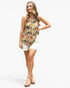 A woman modeling a sleeveless halter-neck top with a colorful tropical floral print, paired with white shorts and tan platform sandals, against a plain light gray background.