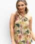 A woman wearing a sleeveless halter-neck dress with a colorful tropical floral and leaf print on a light cream background, photographed against a plain light gray studio background.