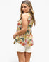 A woman wearing a sleeveless tropical floral print top and white shorts, photographed against a plain light gray studio background.