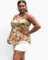 A plus-size model wearing a halter-neck top with a colorful tropical floral and leaf print on a cream background, paired with white shorts, against a plain light gray studio background.
