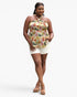 A woman modeling a sleeveless halter-style top with a colorful tropical floral print on a cream background, paired with white shorts and tan platform sandals, against a light gray studio background.
