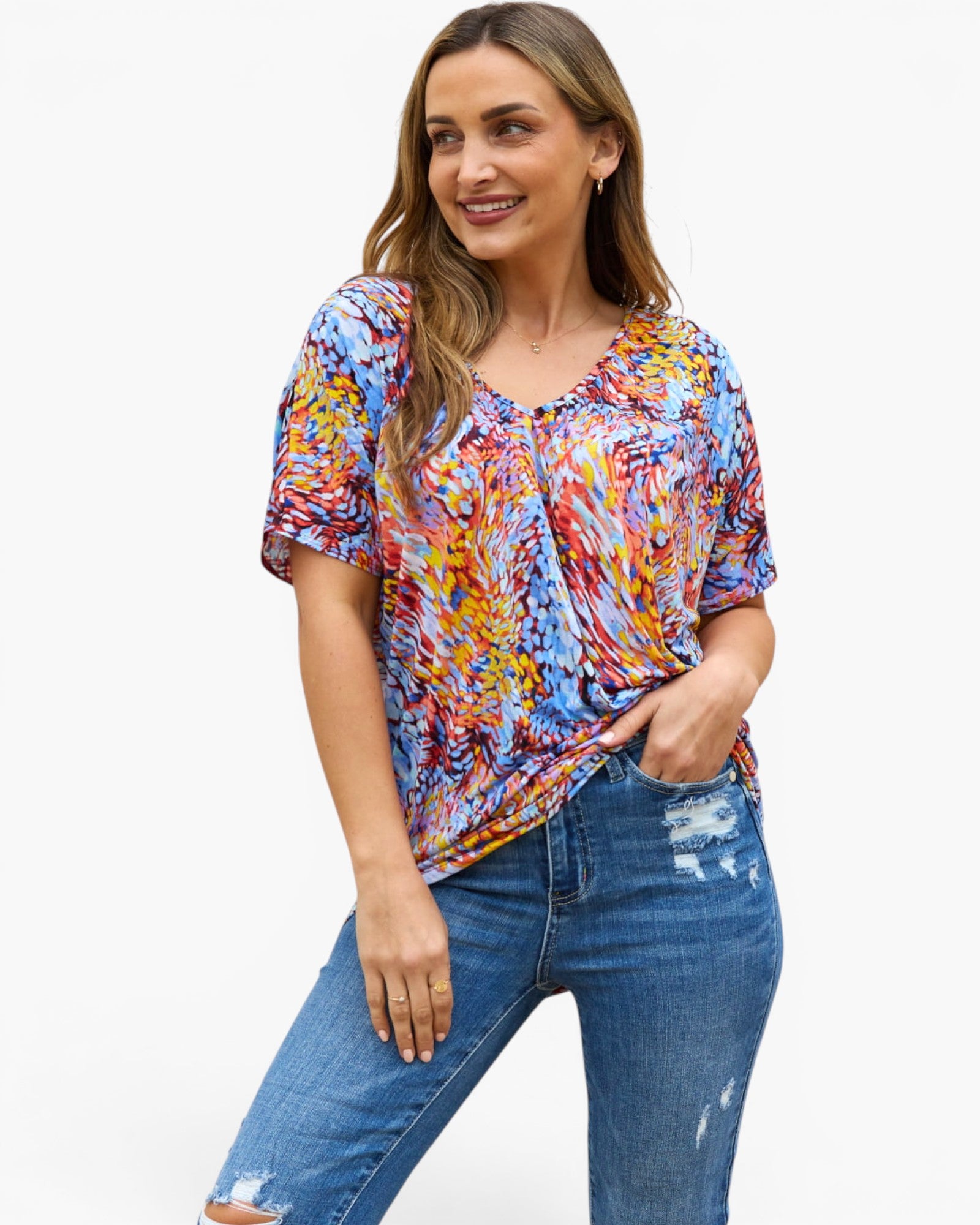 A woman modeling a short-sleeve V-neck top with a vibrant multicolor abstract print, paired with blue distressed jeans, against a plain white background.