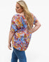 A woman wearing a short-sleeve, loose-fitting top with a vibrant multicolor abstract print, paired with blue jeans, photographed against a plain white background.