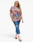 A woman modeling a short-sleeve V-neck top with a vibrant multicolor abstract print, paired with blue jeans and tan sandals, against a plain white background.