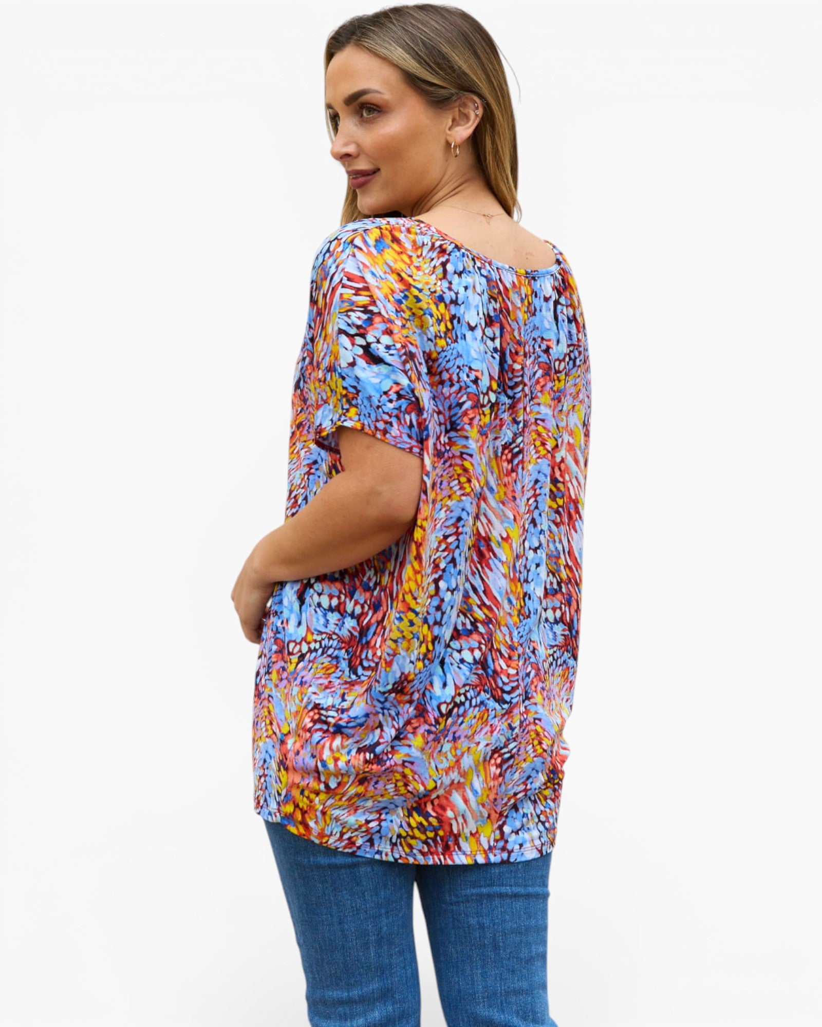 A woman wearing a colorful abstract print short-sleeve top and blue jeans, shown from a three-quarters back view against a plain white background.