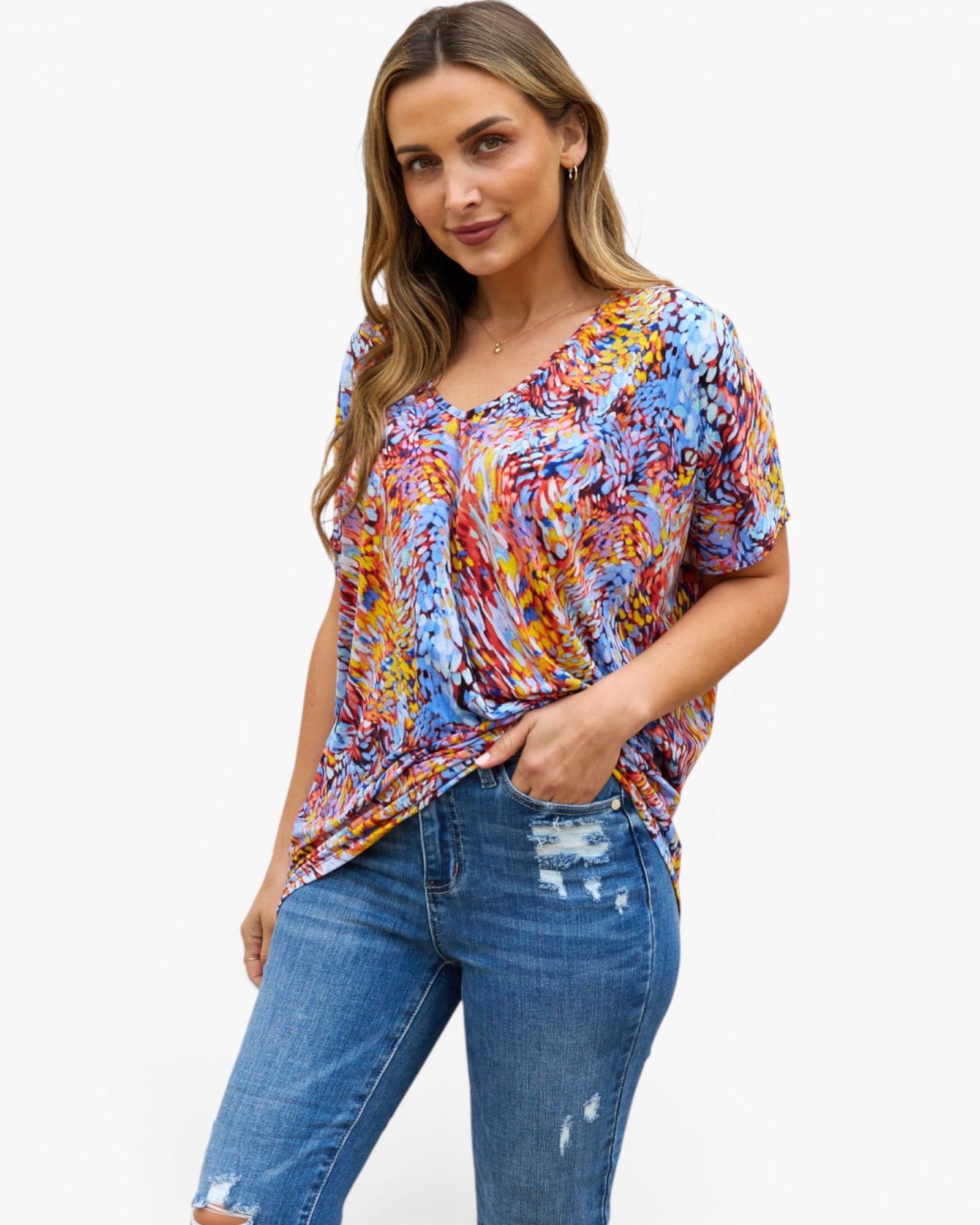 A woman modeling a short-sleeve V-neck top with a vibrant multicolor abstract swirl pattern in blue, red, yellow, and orange, paired with distressed blue jeans against a plain white background.
