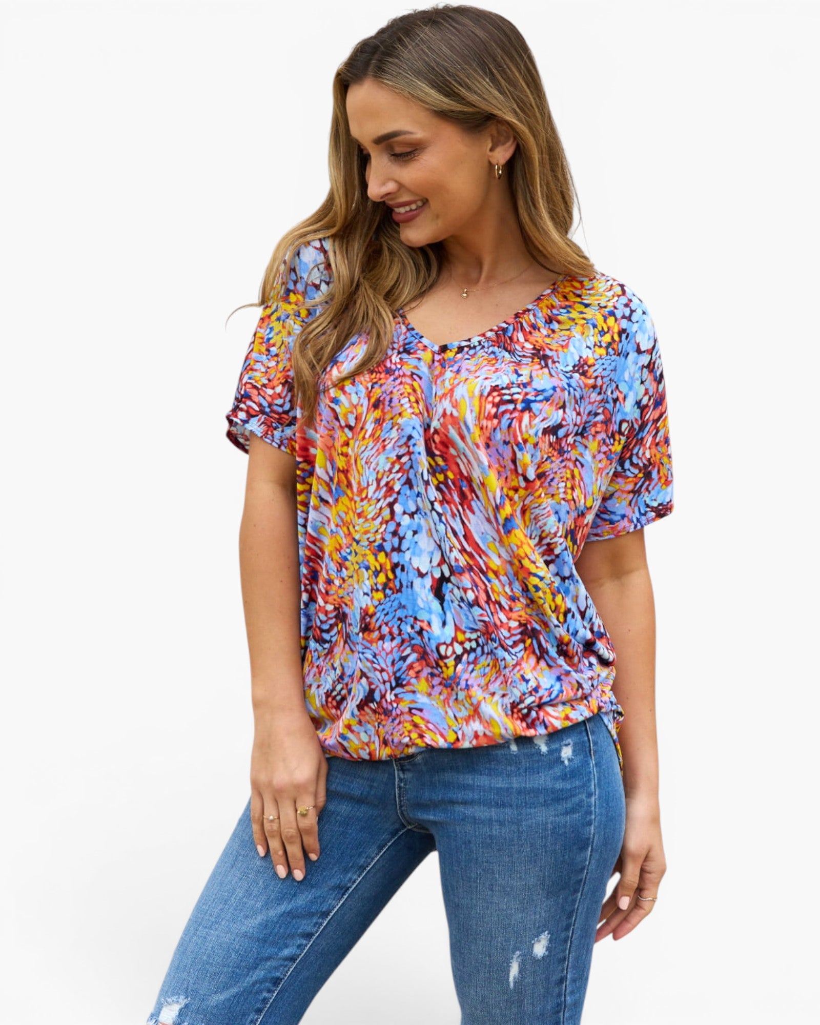 A woman wearing a short-sleeve V-neck top with a vibrant multicolor abstract print in shades of blue, red, yellow, and orange, paired with blue jeans, against a plain white background.