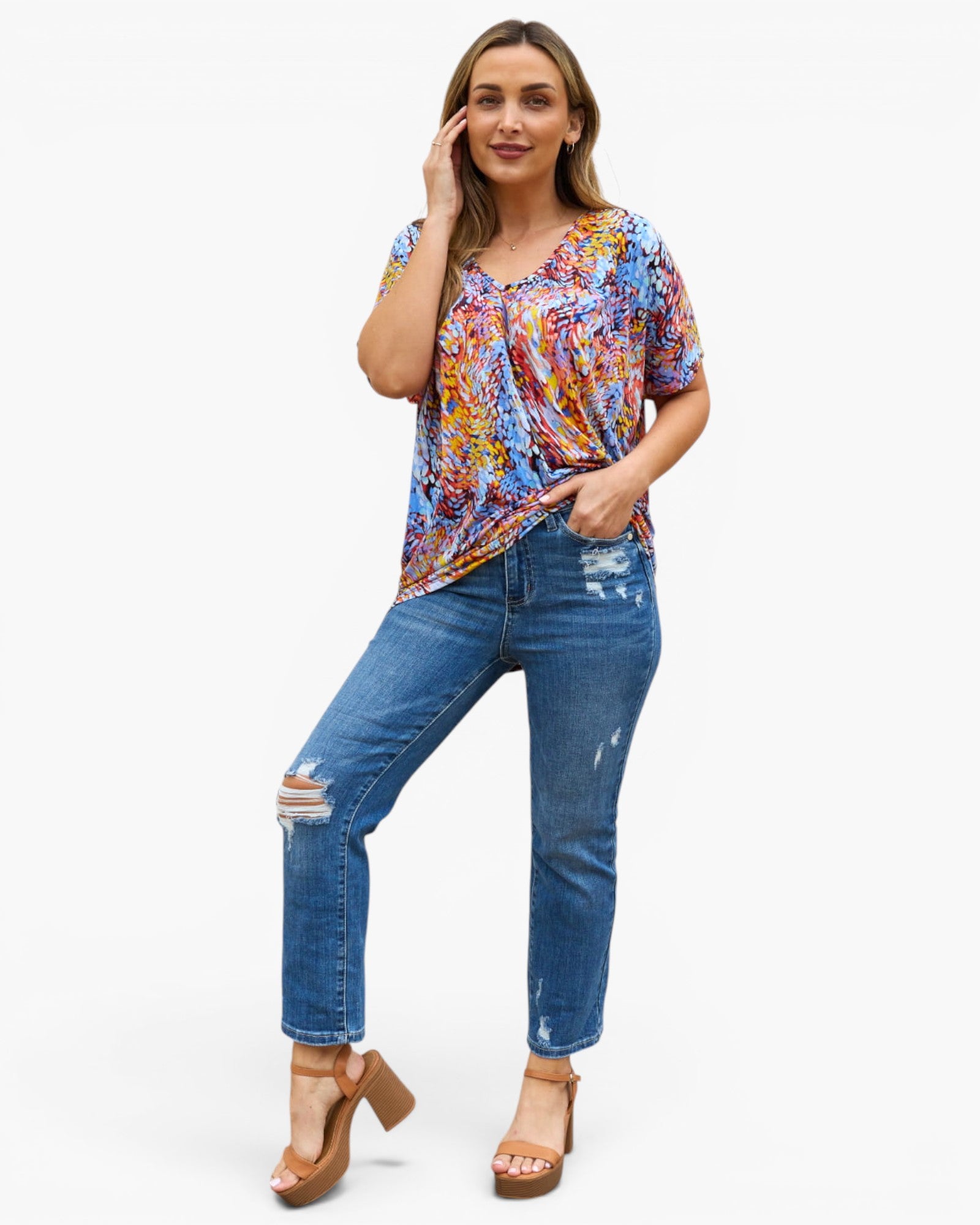 A woman modeling a short-sleeve V-neck top with a colorful abstract print in blue, orange, yellow, and red, paired with distressed blue jeans and brown platform sandals, against a plain white background.