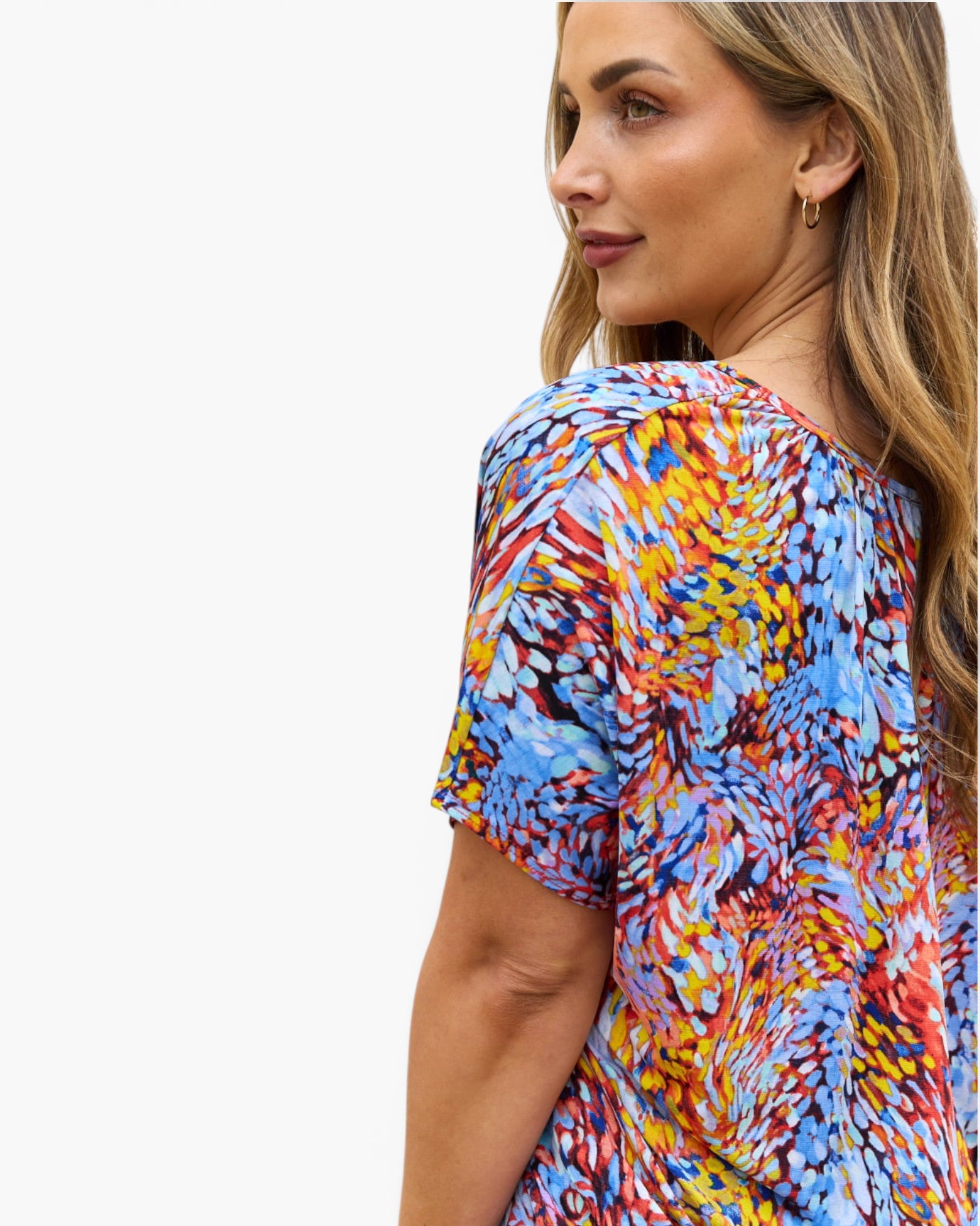A woman wearing a short-sleeve top with a vibrant multicolor abstract brushstroke pattern in blue, red, yellow, and orange, shown from a side-back profile against a plain white background.