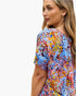 A woman wearing a short-sleeve top with a vibrant multicolor abstract brushstroke pattern in blue, red, yellow, and orange, shown from a side-back profile against a plain white background.