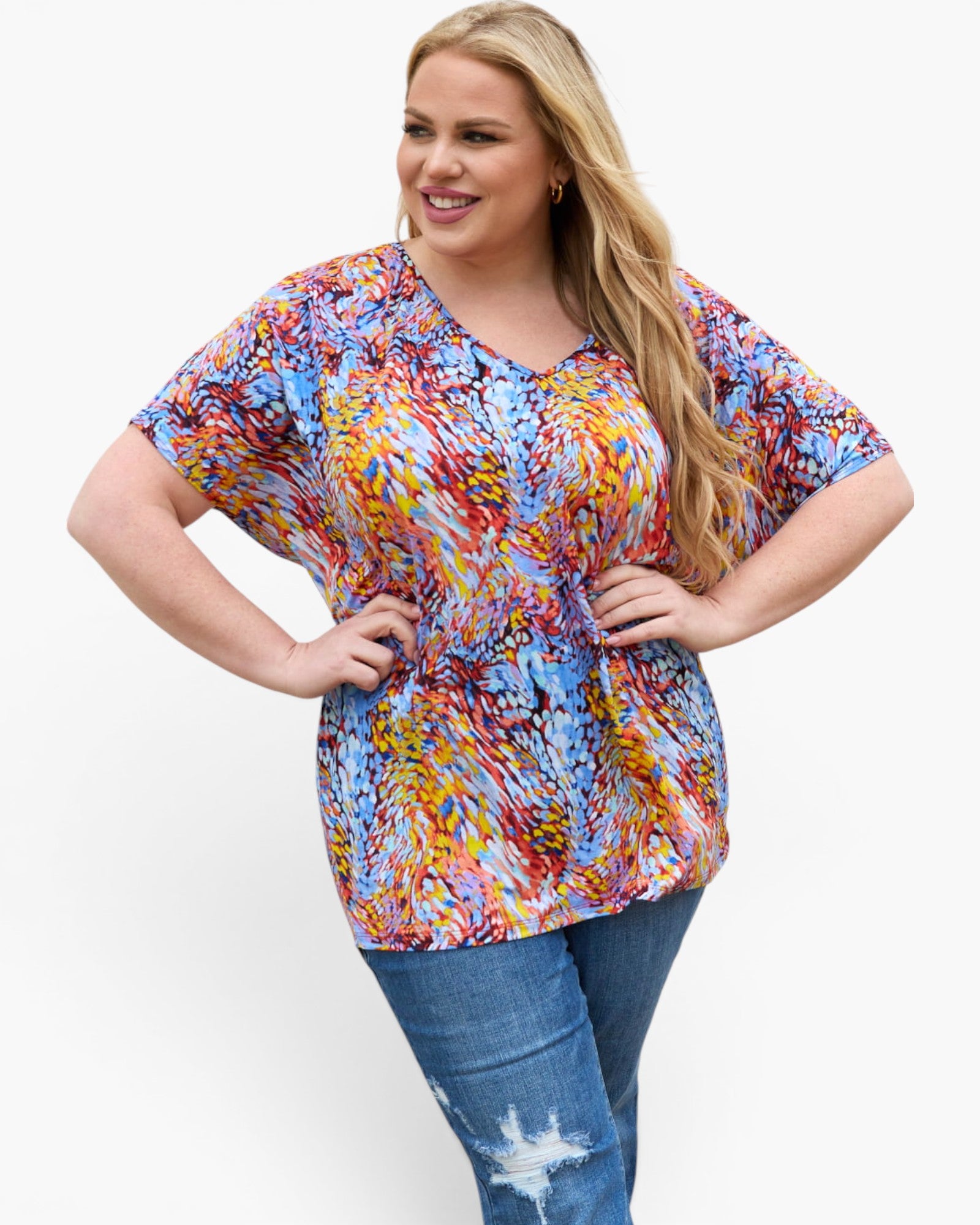 A woman modeling a short-sleeve V-neck top with a colorful abstract brushstroke pattern in shades of blue, red, yellow, orange, and purple, paired with blue jeans against a plain white background.