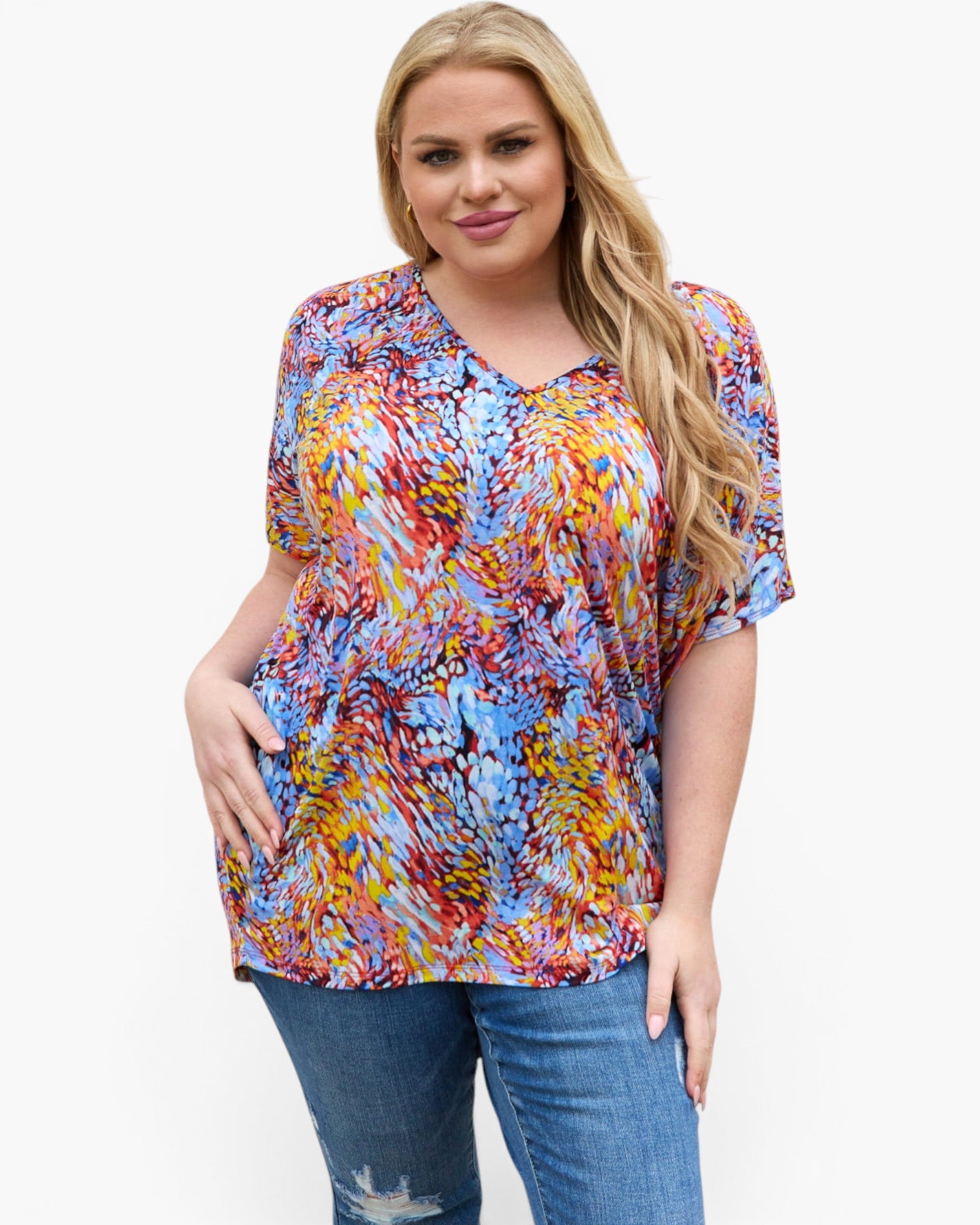 A woman modeling a short-sleeve V-neck blouse with a colorful abstract brushstroke pattern in blue, red, yellow, orange, and white, paired with blue jeans against a plain white background.