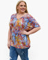 A woman modeling a short-sleeve V-neck top with a colorful abstract brushstroke pattern in blue, red, yellow, orange, and white, paired with blue jeans against a plain white background.