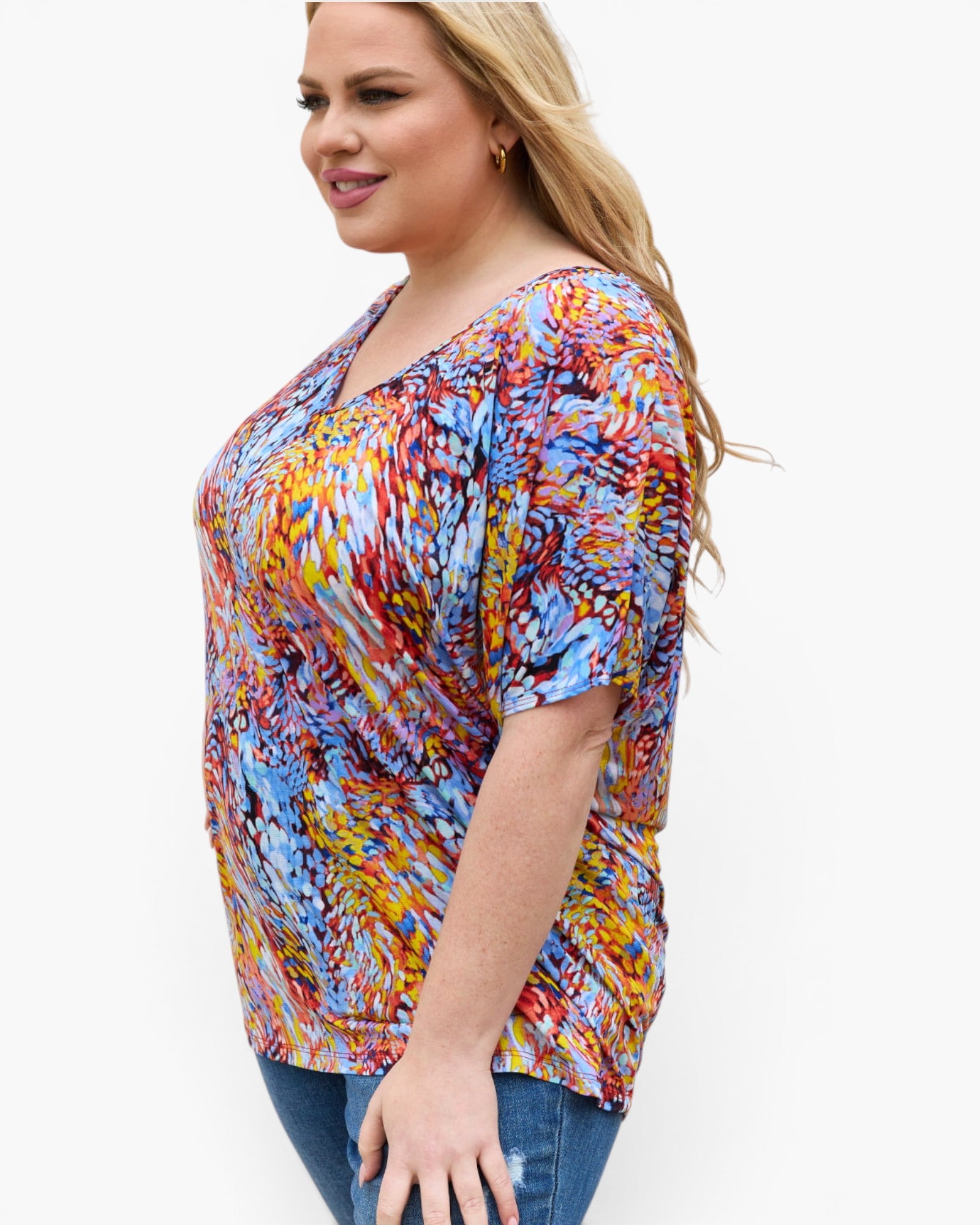 A woman wearing a short-sleeve V-neck top with a colorful abstract brushstroke pattern in blue, yellow, orange, red, and white, paired with blue jeans, shown against a plain white background.