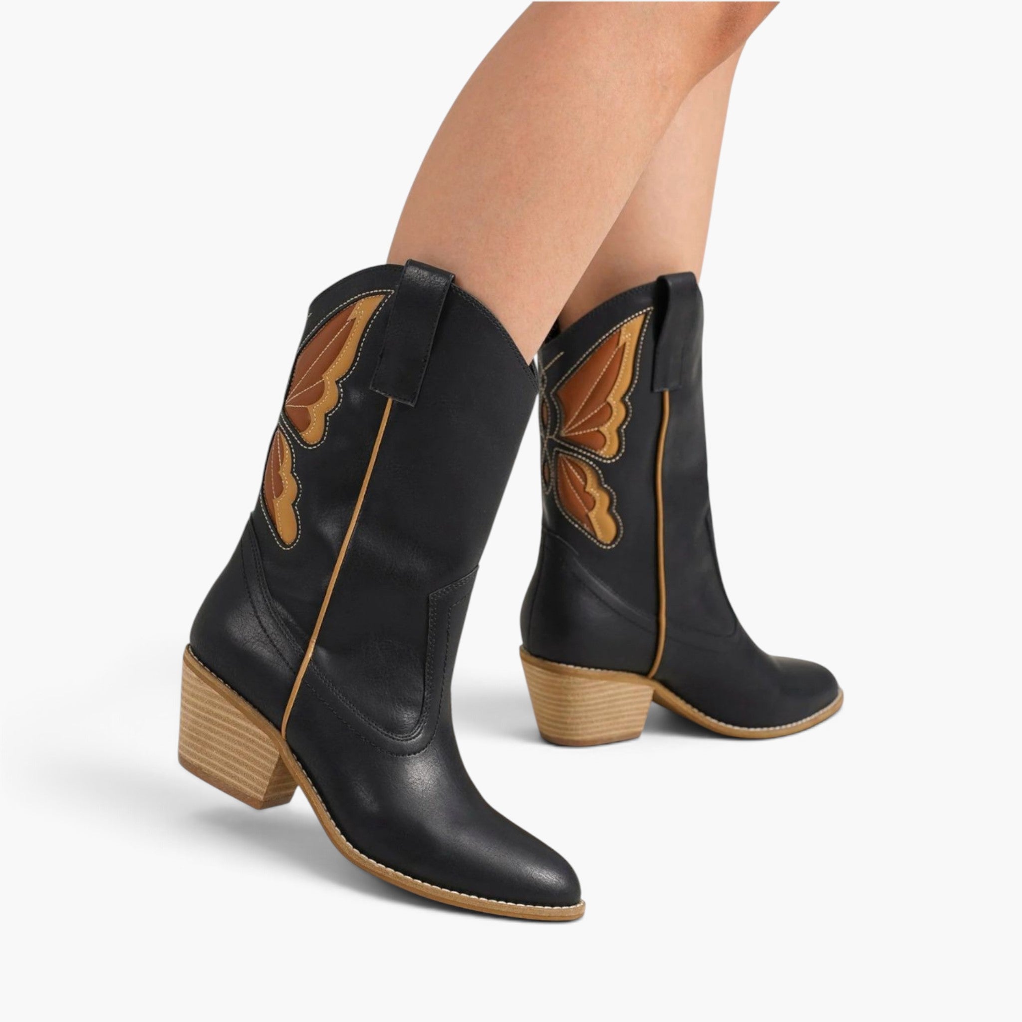 A pair of black leather cowboy boots with brown and orange butterfly wing embroidery on the shaft and tan block heels, shown on a model's feet against a light gray background.