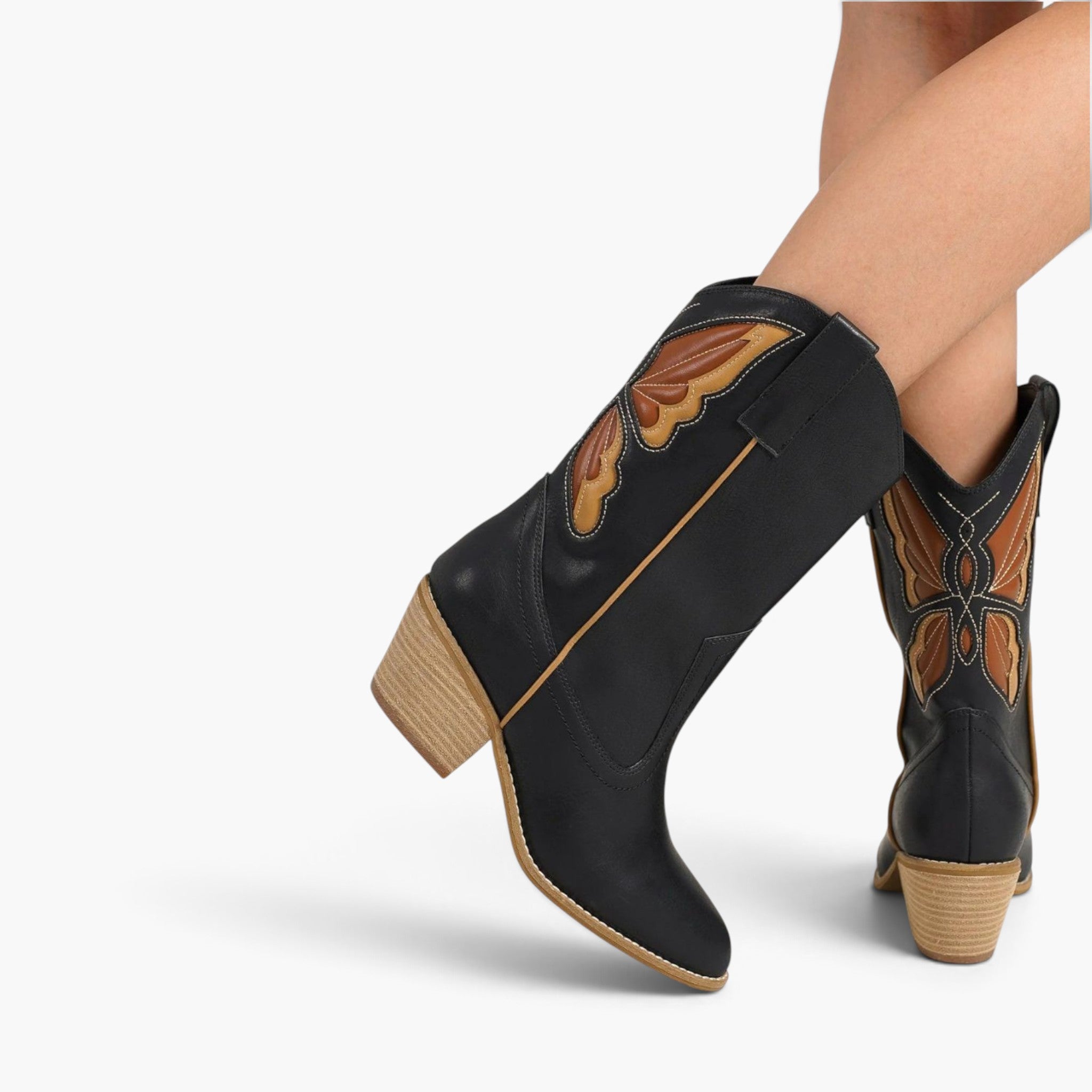Black faux-leather western ankle boots with brown and tan embroidered butterfly details and stacked tan block heels, shown on a model's feet against a light gray studio background.