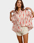 Woman wearing a white blouse with red patterns and denim shorts on a plain background