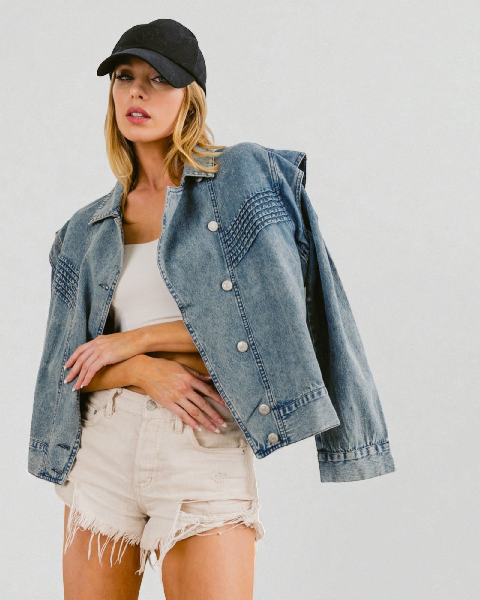 Woman wearing a denim jacket, white top, and shorts with a black cap on a plain background