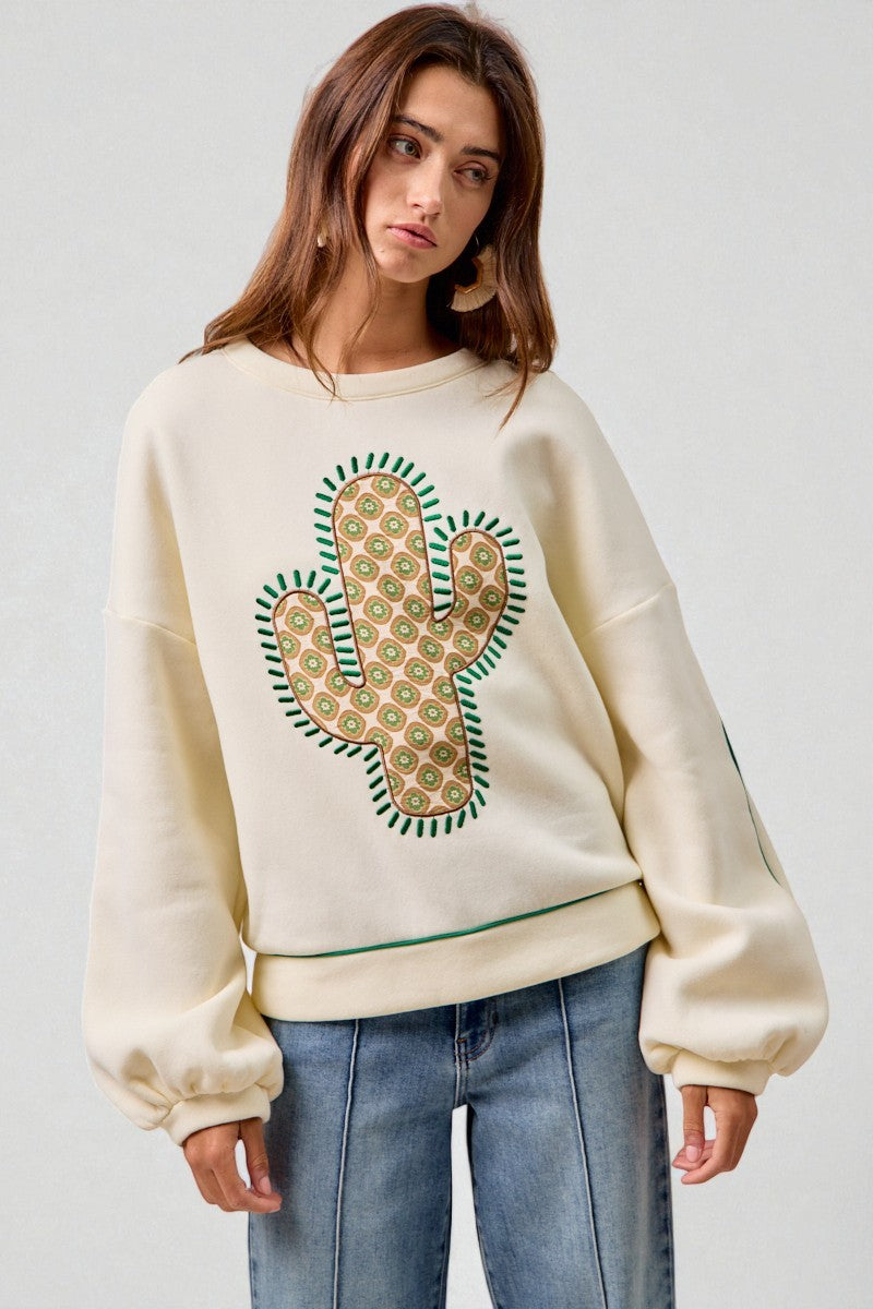 Woman wearing a cream sweatshirt with a cactus design on a plain background