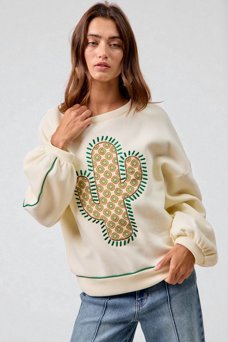 Woman wearing a cream sweatshirt with a cactus design on a plain background