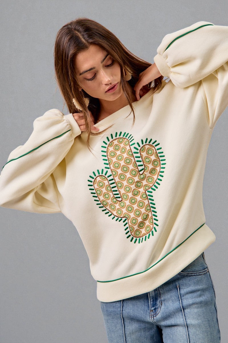 Woman wearing a cream sweatshirt with a green and brown pattern on a gray background