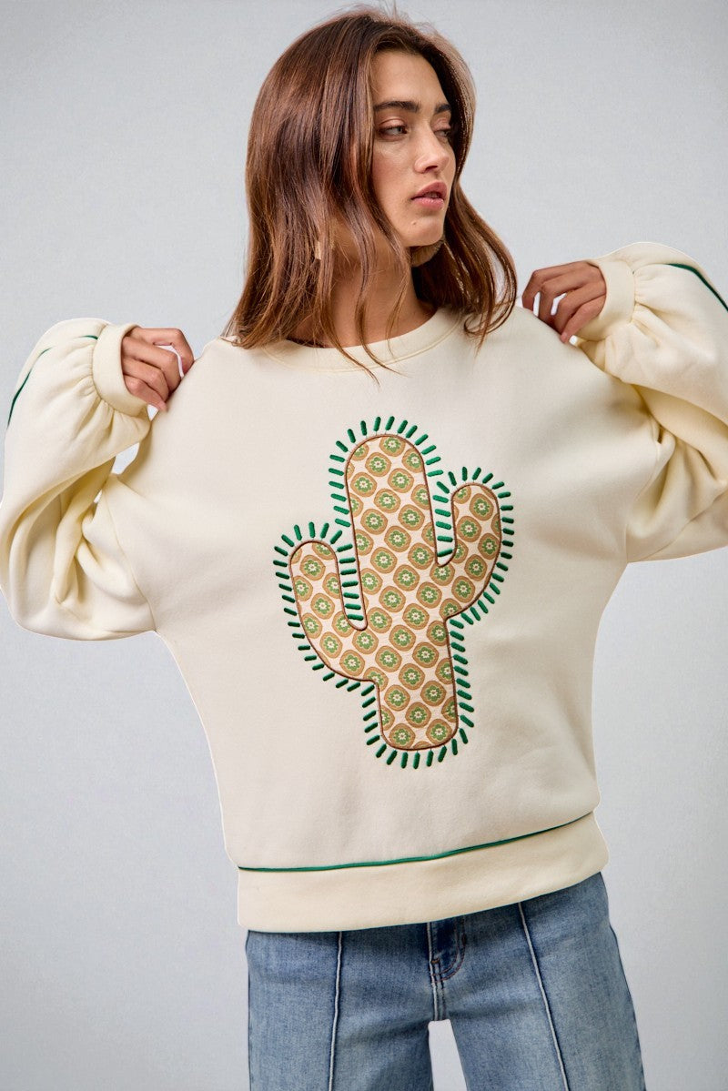 Woman wearing a cream sweatshirt with a cactus design on a plain background
