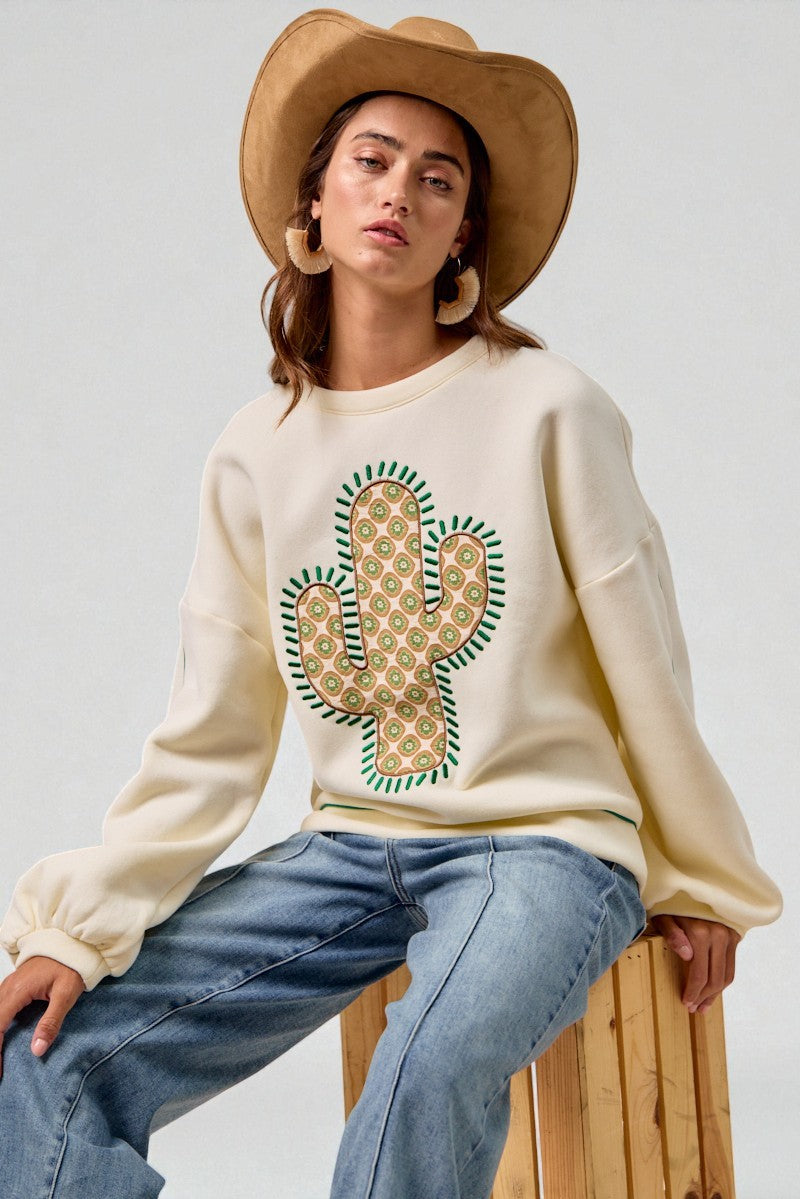 Person wearing a cream sweatshirt with a cactus design, blue jeans, and a beige hat on a white background.