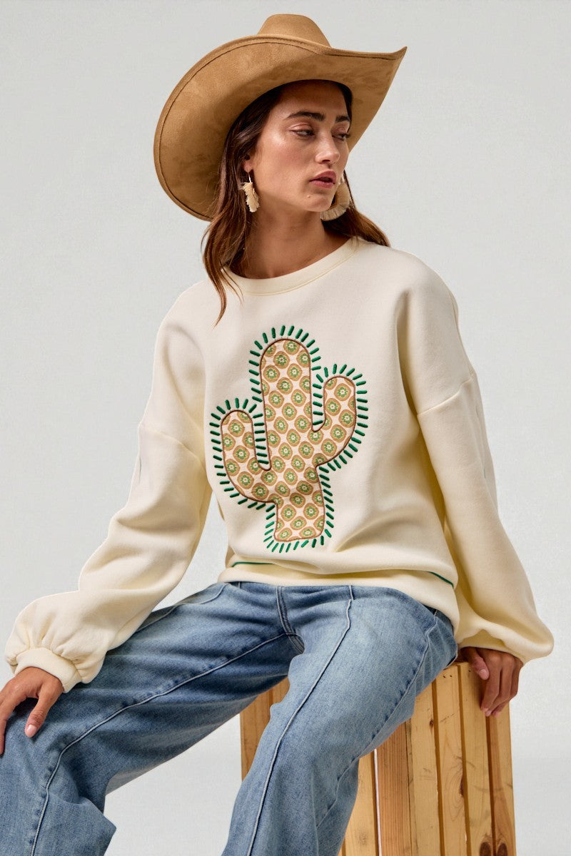 Person wearing a cream sweatshirt with a cactus design, blue jeans, and a brown cowboy hat on a light gray background.
