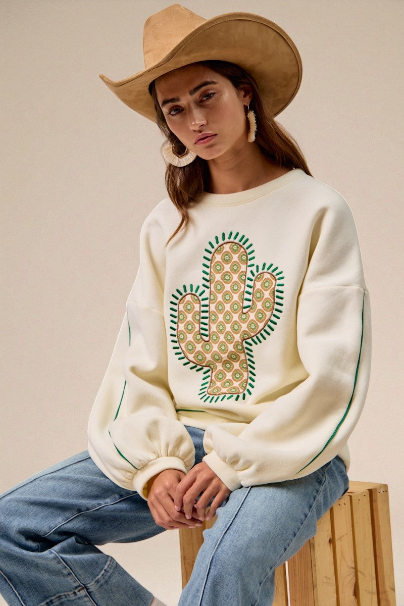 Woman wearing a cream sweatshirt with a cactus design, sitting on a wooden stool.