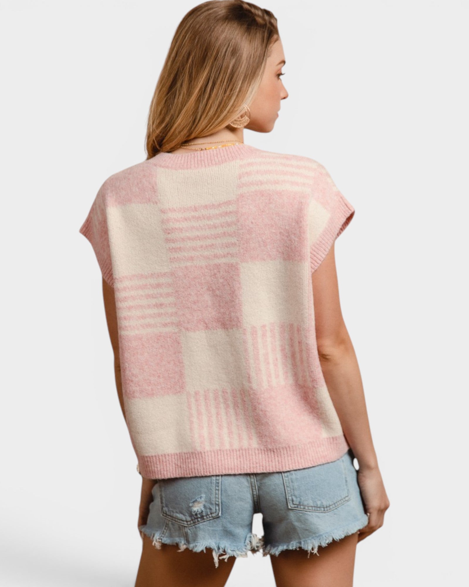 Woman wearing a pink and beige checkered sleeveless sweater with denim shorts on a white background