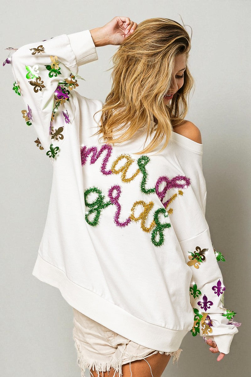 Woman wearing a white sweatshirt with colorful 'Mardi Gras' text and floral designs on a plain background