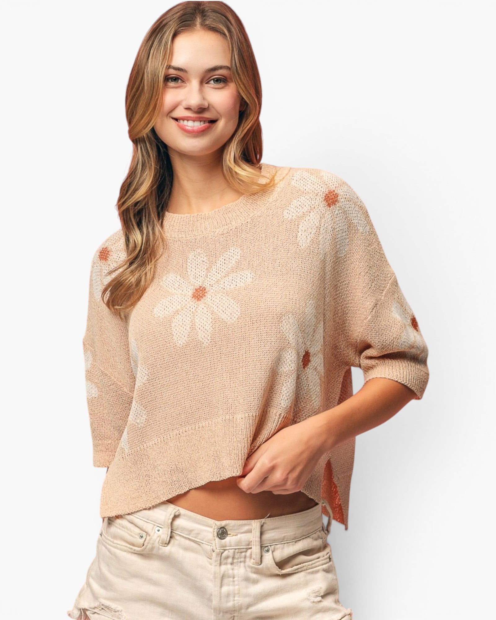 A woman wearing a beige knit sweater with large white daisy patterns and small orange flower centers, paired with light beige jeans, against a plain white background.