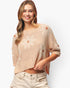 A woman wearing a beige knit sweater with large white daisy patterns and small orange flower centers, paired with light beige jeans, against a plain white background.
