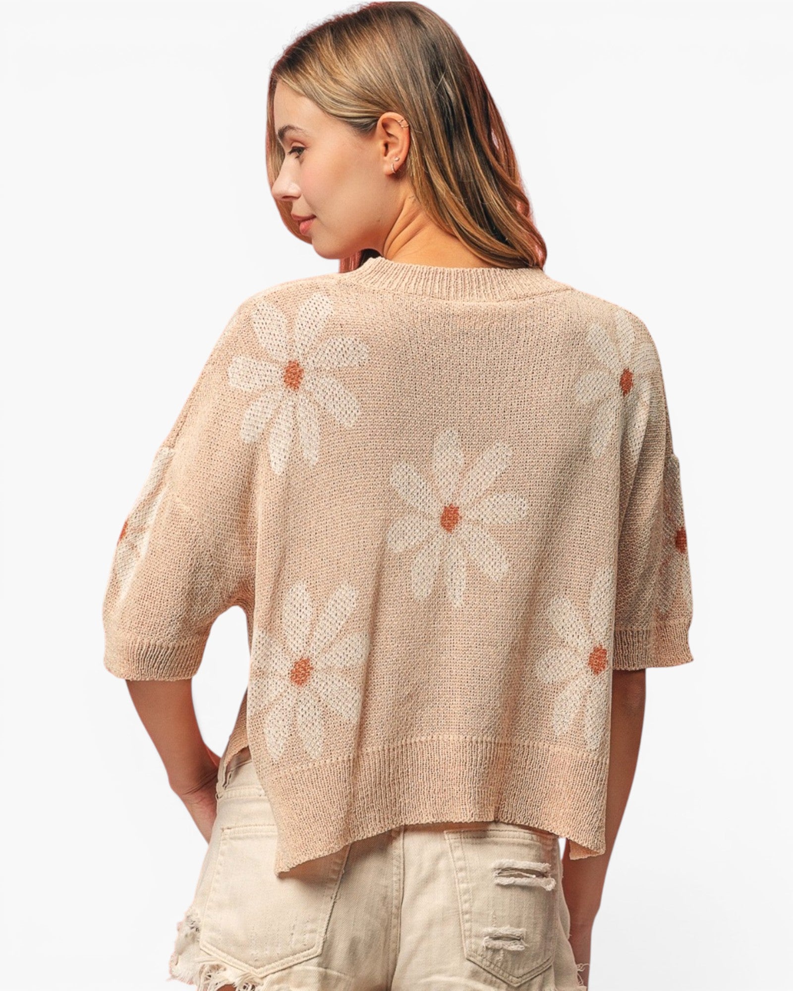 A woman shown from the back wearing a beige knit sweater with large white daisy patterns and tan centers, paired with light beige shorts, against a plain white background.