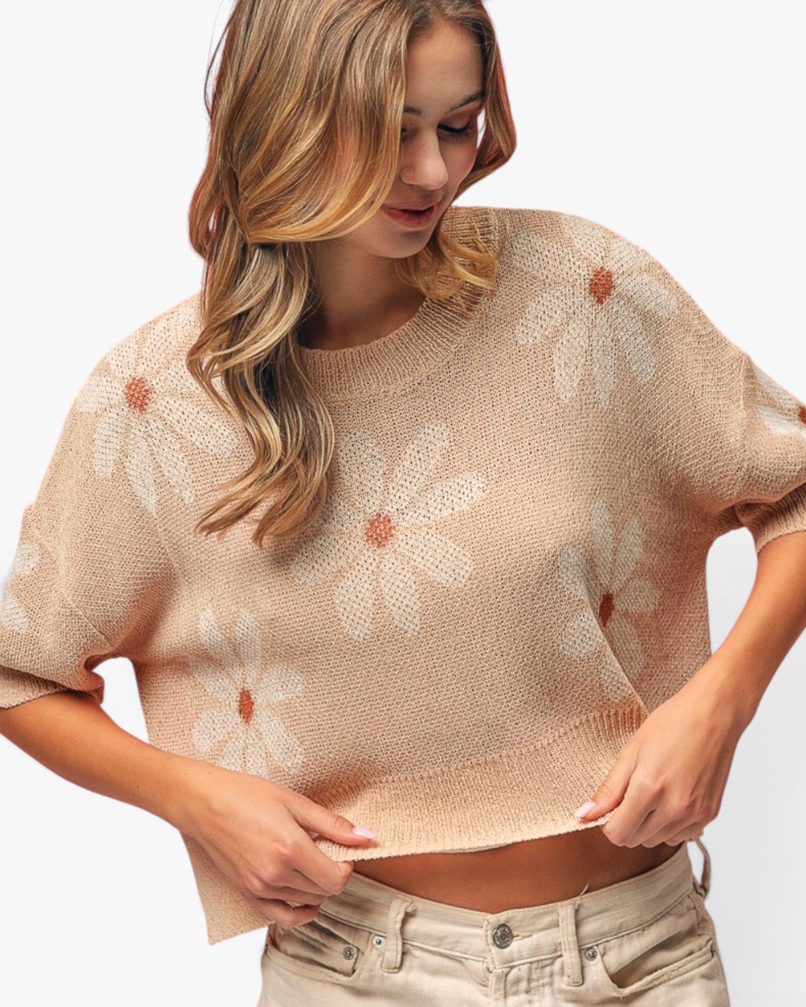 A woman wearing a beige knit short-sleeve crop top with a white daisy pattern and light beige jeans, shown against a plain white background.