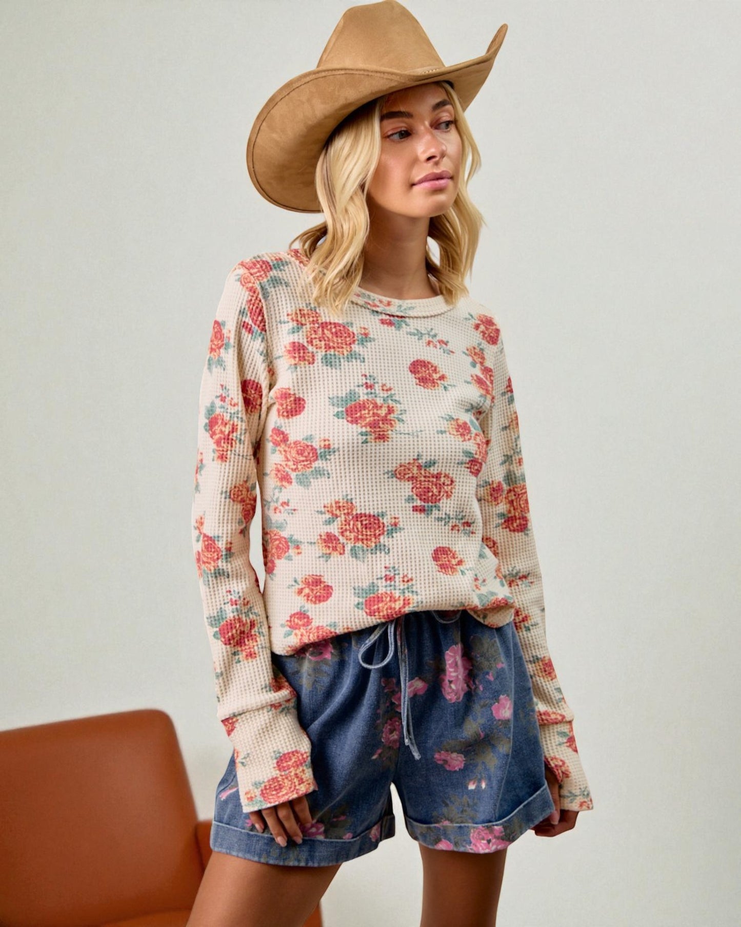 Woman wearing a floral sweater and denim shorts with a brown hat on a plain background